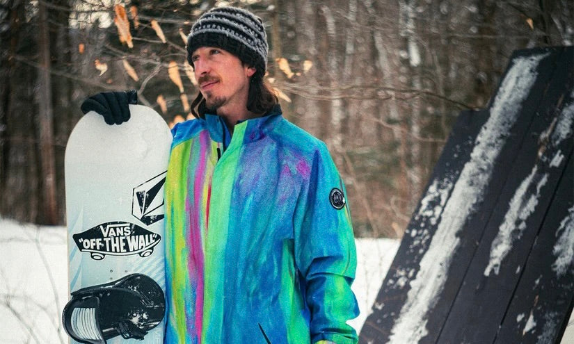 snowboarder with a rainbow coloured jacket and a snowboard stood in amongst snowy trees