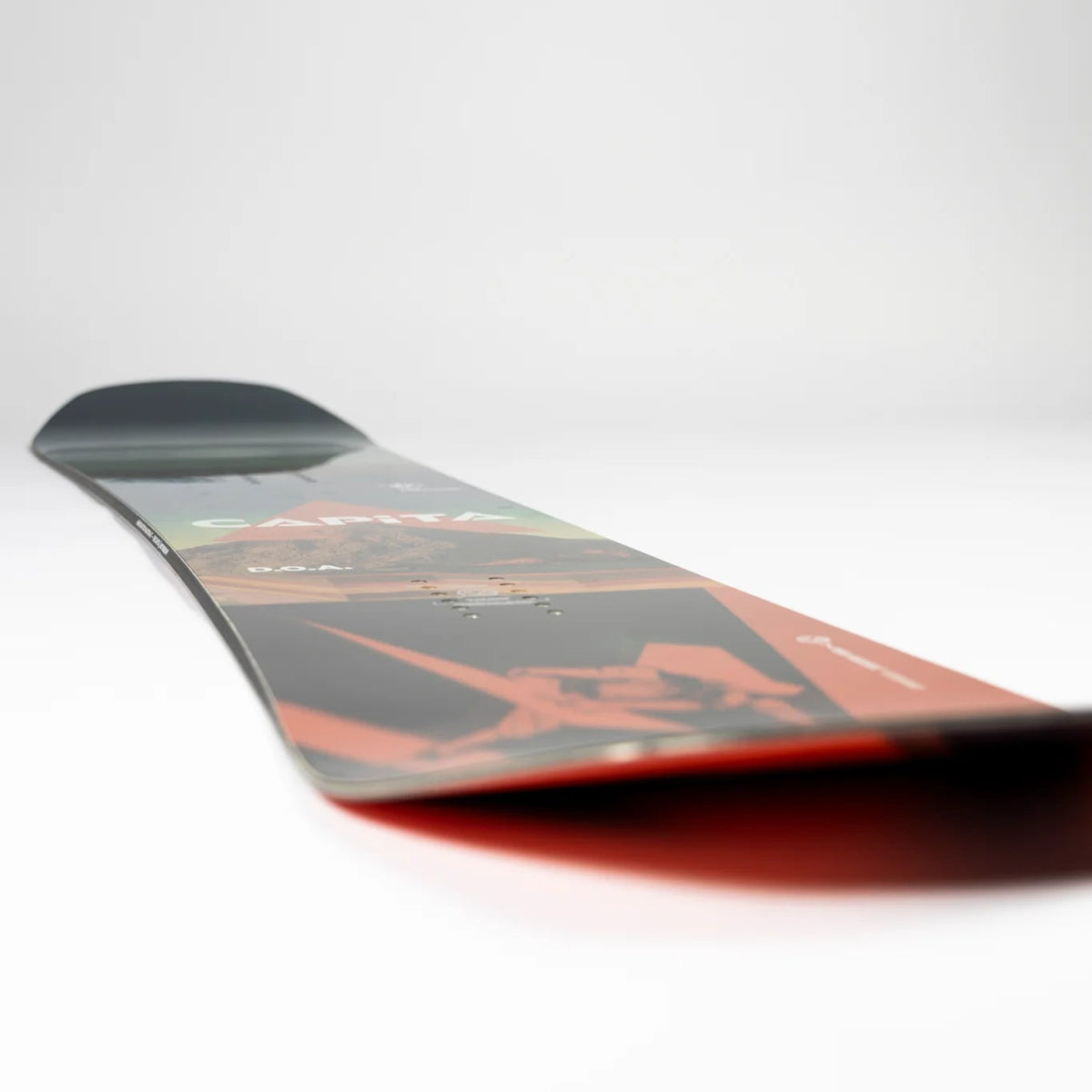 A close-up, angled view of a snowboard's base showing a dark gray and black graphic pattern with a prominent orange-red accent line and a red underside, set against a clean white studio background.
