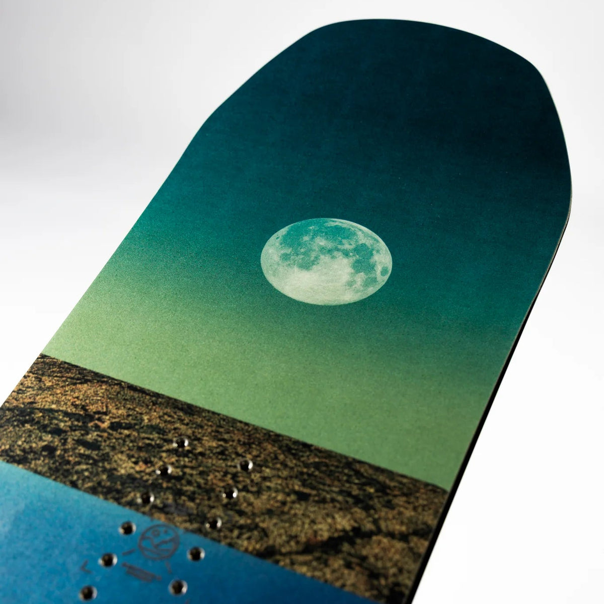 A close-up of a snowboard deck featuring a scenic graphic print of a full moon over a landscape, with a teal-to-blue gradient and a textured brown horizon.