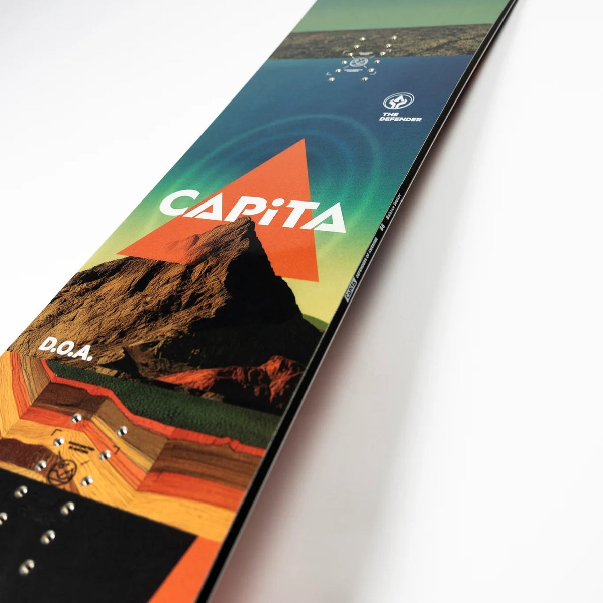 A single snowboard photographed on a clean white studio background, shown at an angle with the top graphic side facing up. The board features a scenic print with a mountain, water, sky gradient, and large orange CAPiTA logo text.