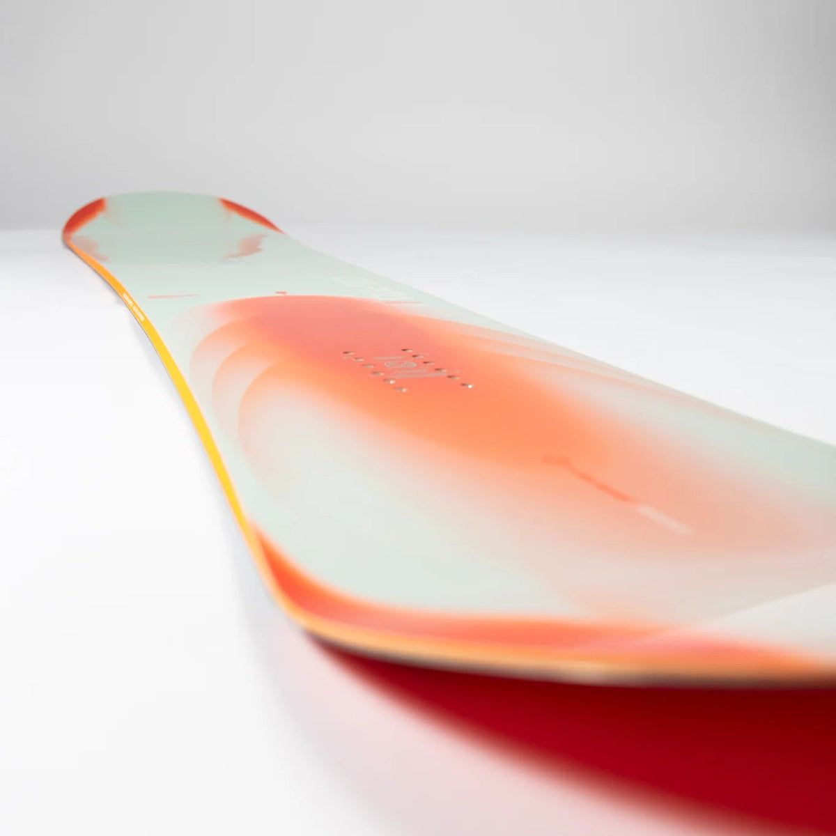 A close-up studio shot of a snowboard deck showing its base with a gradient design transitioning from pale mint green to vibrant coral orange and deep red, with a thin yellow edge.
