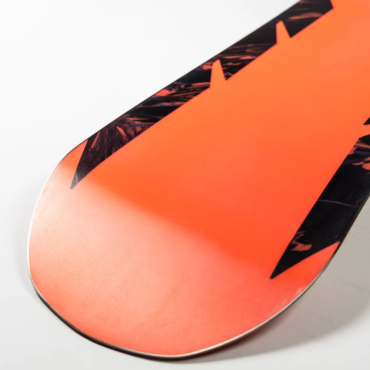 Close-up of the tail section of a snowboard with a bright orange base and black graphic pattern, photographed on a light gray background.