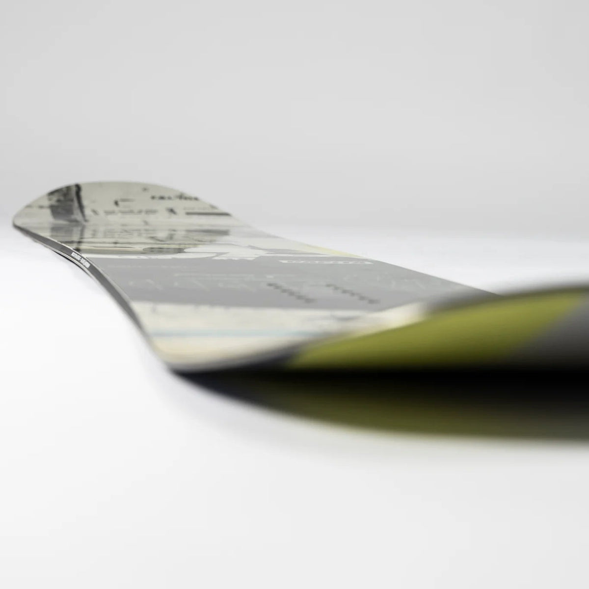 A close-up, angled view of a snowboard featuring a black, gray, and white graphic pattern with a dark olive green base on a clean white background.