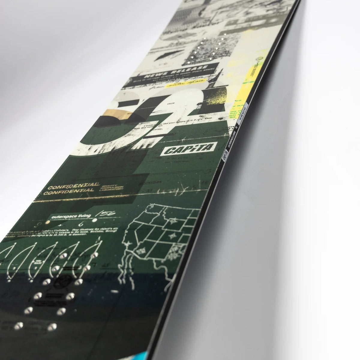 A close-up angled view of a snowboard deck featuring a complex graphic design with black, white, green, yellow, and blue elements, including the CAPiTA logo.