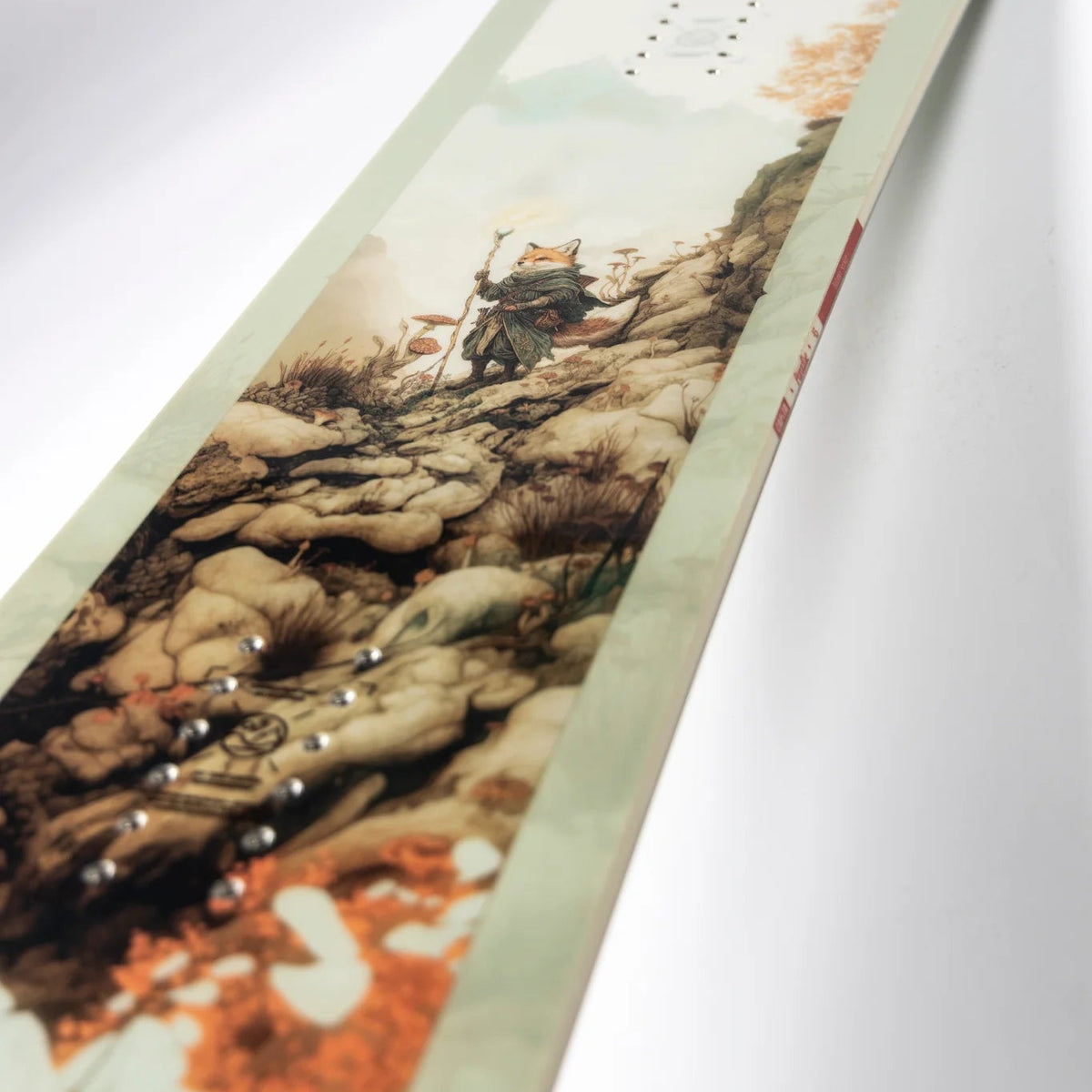 A close-up of a snowboard deck featuring a scenic graphic print of a mountain climber on rocky terrain, with a pale sage green background and orange autumn foliage accents.