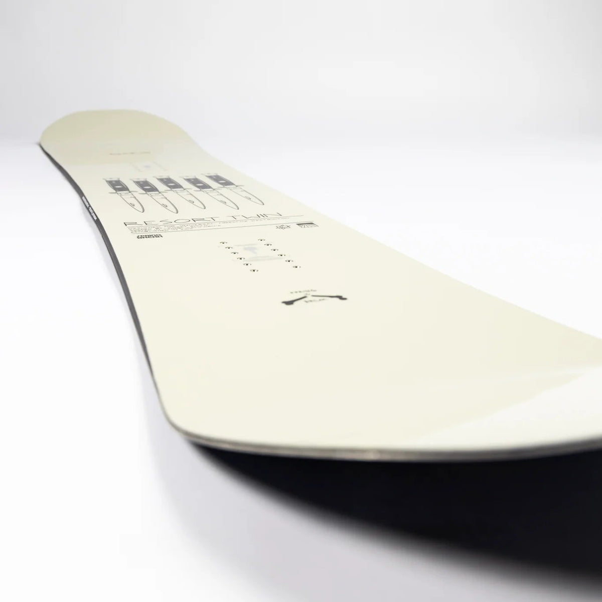 A close-up, angled view of a snowboard's base showing the cream-colored surface with black printed graphics and binding track, set against a plain white background.