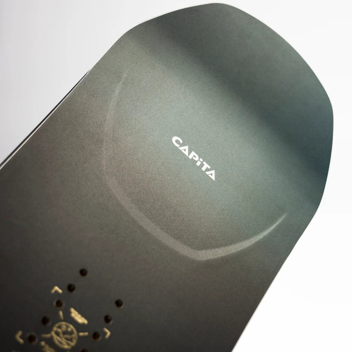 Close-up of a snowboard tail section with the brand name CAPiTA visible in white text, showing a dark gray to black finish with a small gold logo and a hint of orange graphic near the bottom left.