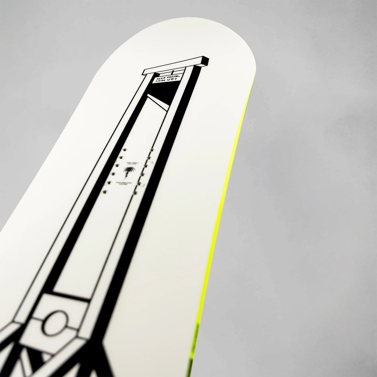 A close-up of a white snowboard featuring a black geometric graphic of a guillotine and a bright neon yellow edge, set against a neutral gray background.