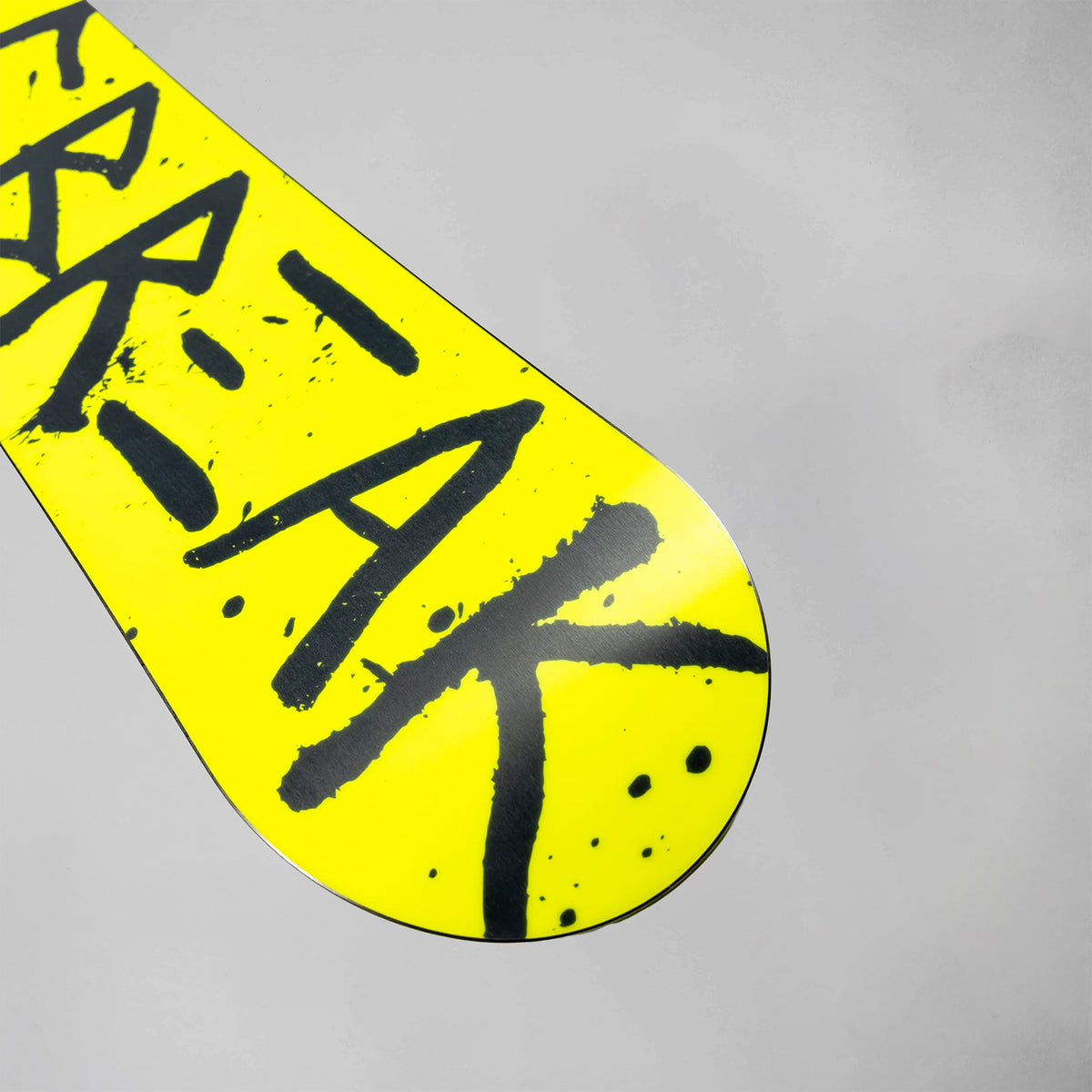 A close-up of a bright yellow snowboard tail with bold black graffiti-style lettering, photographed against a plain light gray background.